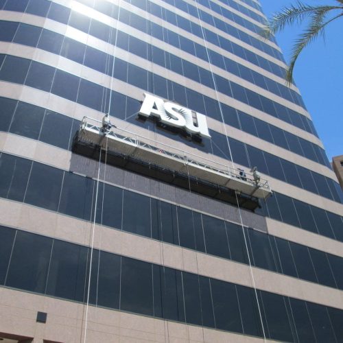 A window-washing platform is suspended in front of a high-rise building with an "ASU" sign under a clear blue sky.