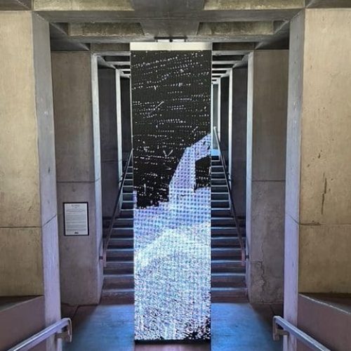 A tall, rectangular art installation with a pixelated black and white design stands at the base of a concrete staircase in an industrial-style building.