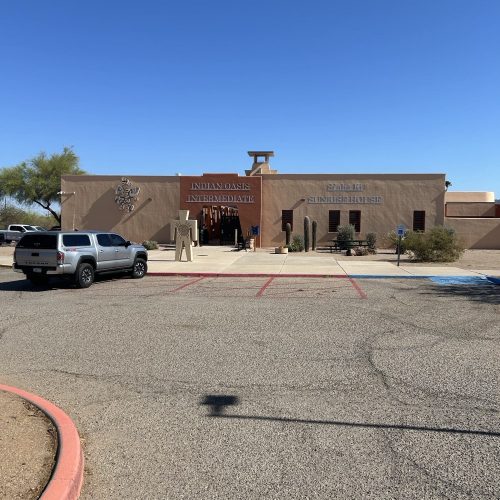 A beige, single-story building labeled "Indian Oasis Intermediate" and "S'bgadig Summer House" with two parked vehicles in front under a clear blue sky.