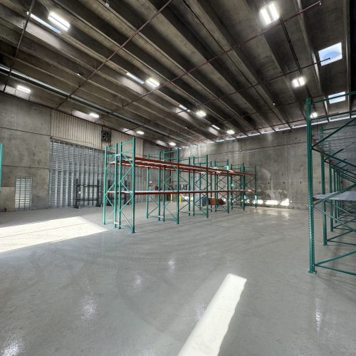 Large, mostly empty warehouse interior with tall concrete walls, overhead lighting, and several green metal storage racks lined up along the polished floor.