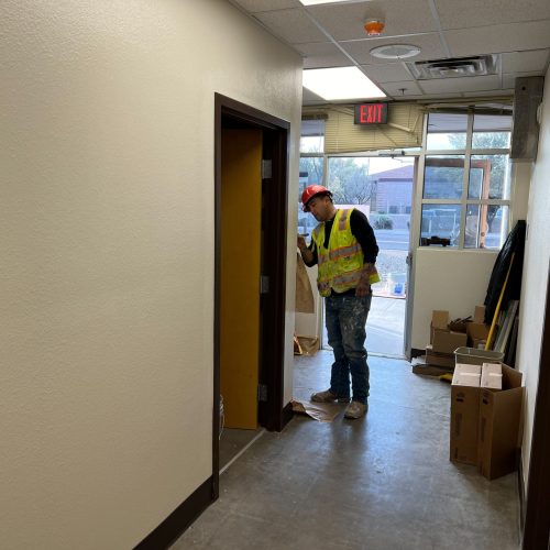 A construction worker wearing a safety vest and hard hat inspects a doorway in a hallway under renovation, with boxes and tools nearby.
