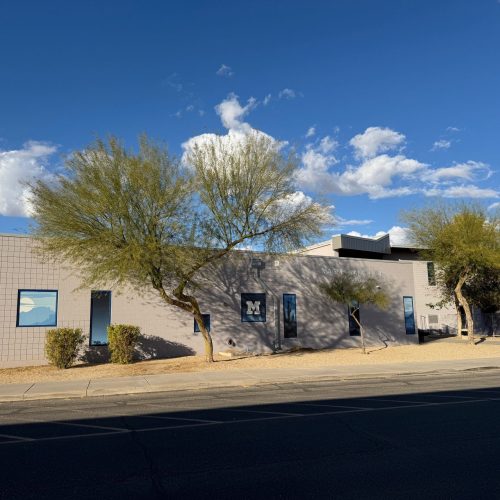 Single-story beige building with a few windows, desert landscaping, and sparse trees under a blue sky with scattered clouds.