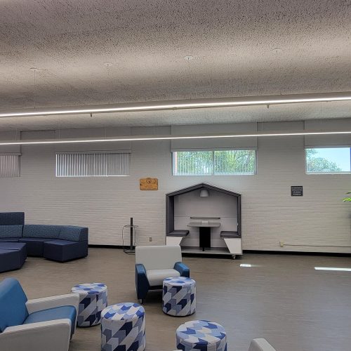 A modern lounge area with blue and white seating, geometric ottomans, a built-in desk nook, and large windows letting in natural light.