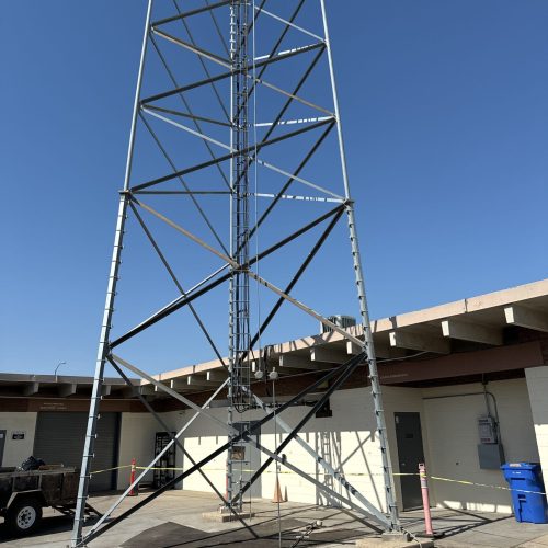 A metal communication tower stands on a concrete base next to a beige building under a clear blue sky; yellow caution tape surrounds the area.