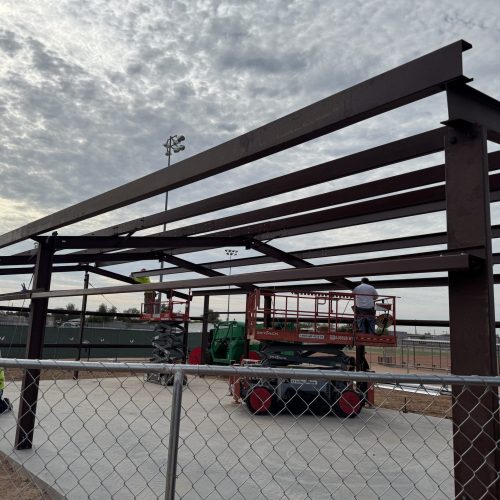 A steel framework for a building under construction, with workers on a lift and others on the ground, surrounded by a chain-link fence on a cloudy day.