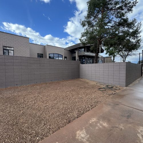 A fenced area with bare dirt ground, coiled hoses, and a concrete path next to a modern building with trees and a partly cloudy sky overhead.