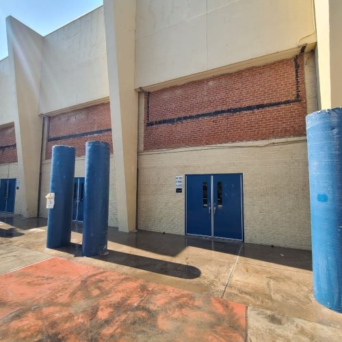 Blue double doors set into a beige and red brick building, with tall blue cylindrical bollards along the sidewalk in front. Sunlight creates shadows on the wet ground.