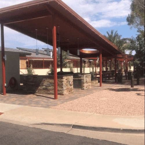 A modern outdoor covered patio area with stone and metal accents, paver flooring, landscaping, and picnic tables under a sloped red roof.