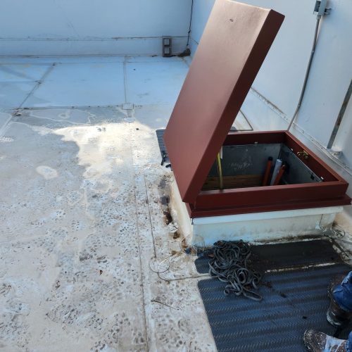 A person in a neon shirt stands near an open rooftop hatch with tools and rope visible beside the hatch.